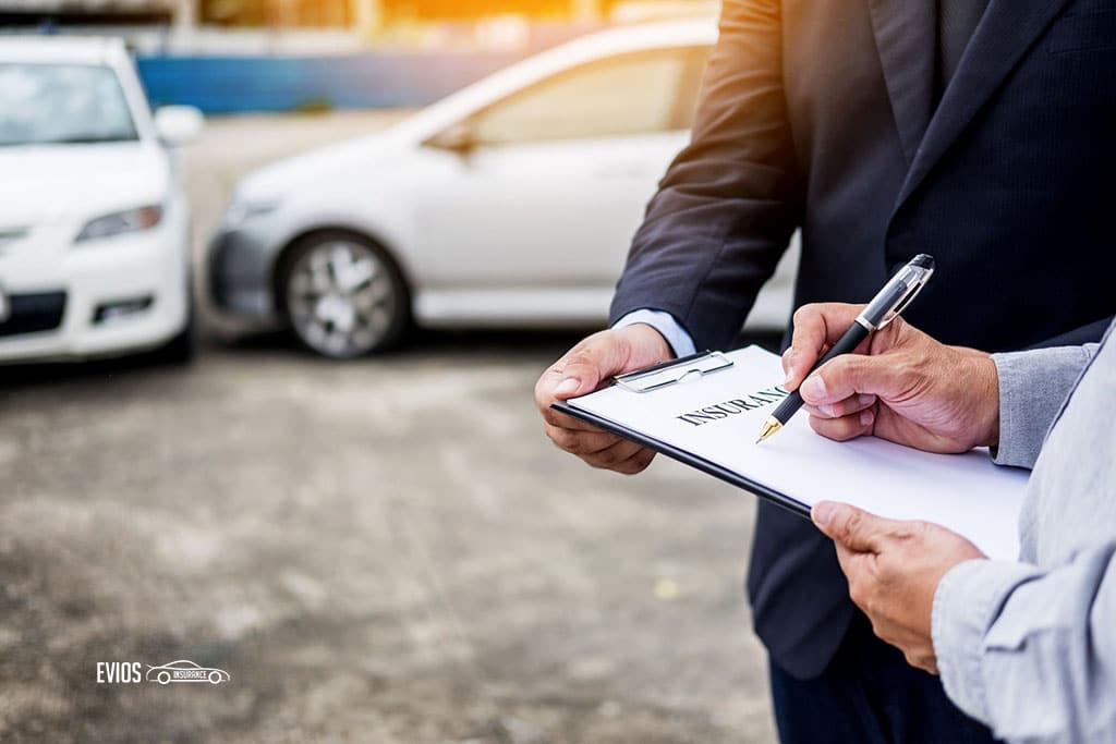  A man in a suit is writing on a clipboard while another man looks over his shoulder. The two men are standing in front of two damaged cars. The image is about third party insurance for car accidents.